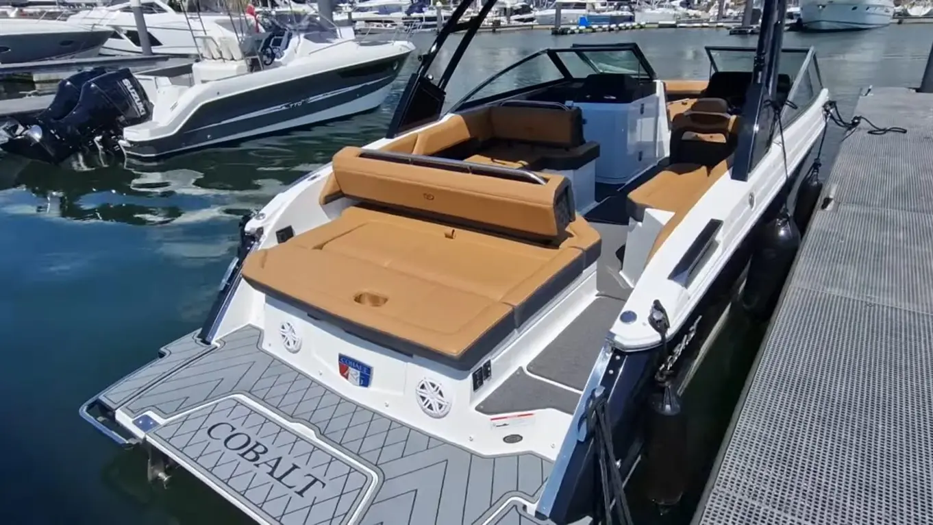 A Cobalt boat with tan cushions is docked by a marina, showcasing its spacious rear seating and sleek design, reflecting in water.